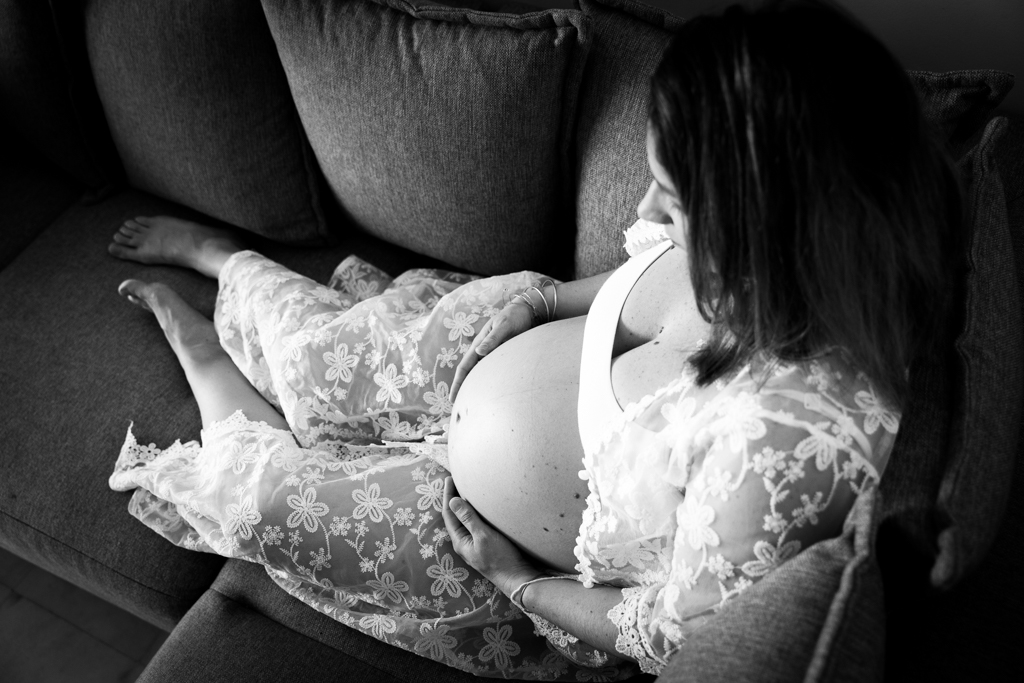 Une femme enceinte pose assise sur son canapé lors d'une séance photo de grossesse à domicile.