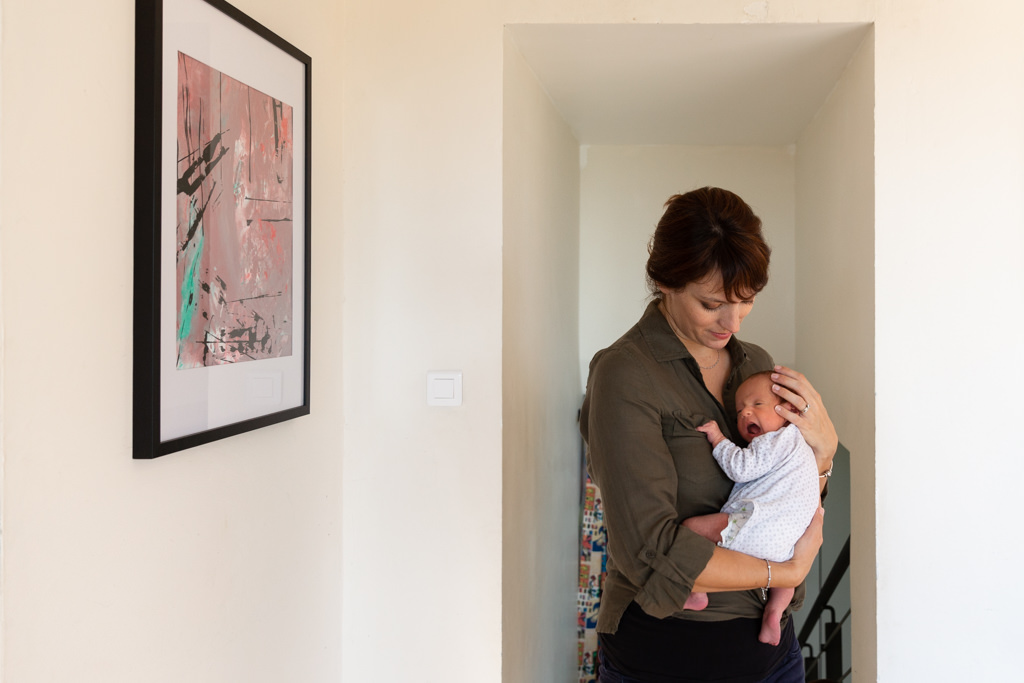 Photo d'une maman debout portant son bébé chez eux. Photo prise à domicile par Myriam Ohayon Photographe au Rouret