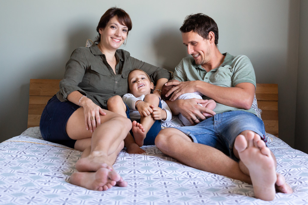 Une famille avec un nouveau né pose sur le lit parental lors d'une séance photo naissance. Photo prise par Myriam Ohayon Photographe au Rouret