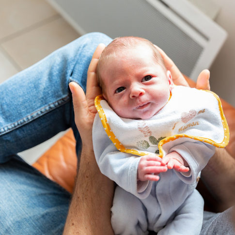 Un bébé est sur les genoux de son papa. Photo prise lors d'une séance photo naissance à Tourrette-Levens