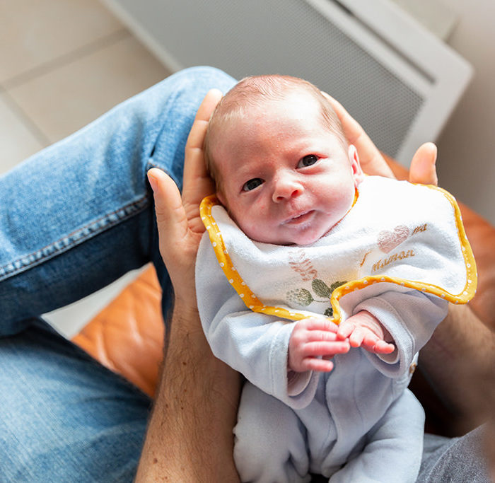 Un bébé est sur les genoux de son papa. Photo prise lors d'une séance photo naissance à Tourrette-Levens