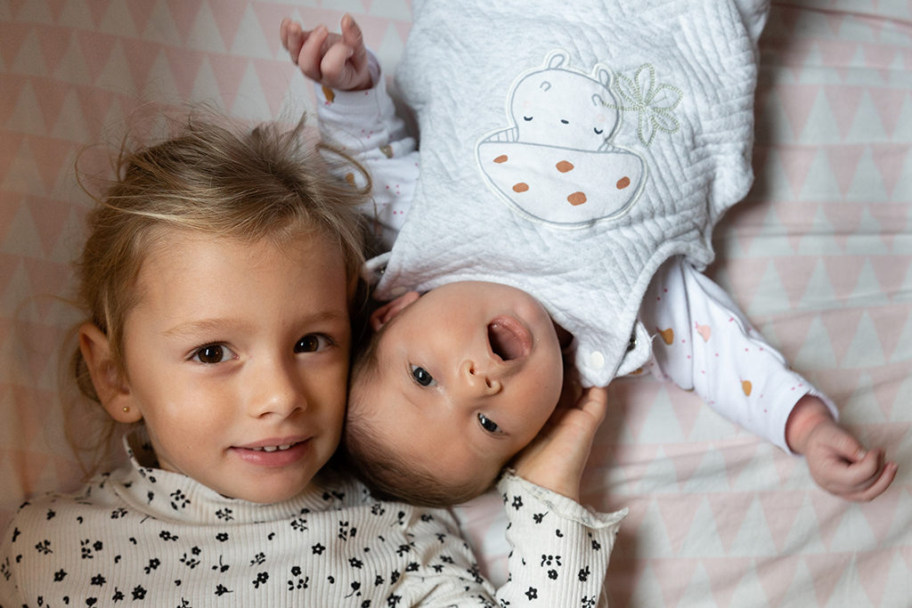 Une grande soeur pose avec sa petite soeur nouveau né lors d'un shooting naissance. Myriam Ohayon Photographe