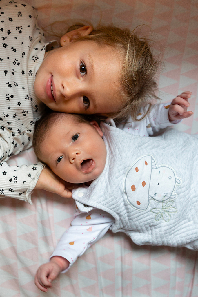 Portrait d'une grande soeur et de sa petite soeur nouveau-né allongées sur le lit. Photo prise.à l'occasion d'une séance photo naissance à Nice