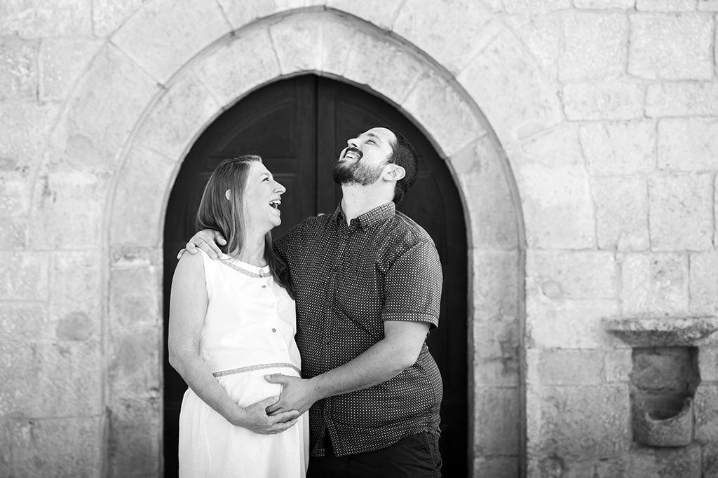 Photo en noir et blanc d'une femme enceinte et de son mari qui posent devant la porte d'une chapelle et éclatent de rire.